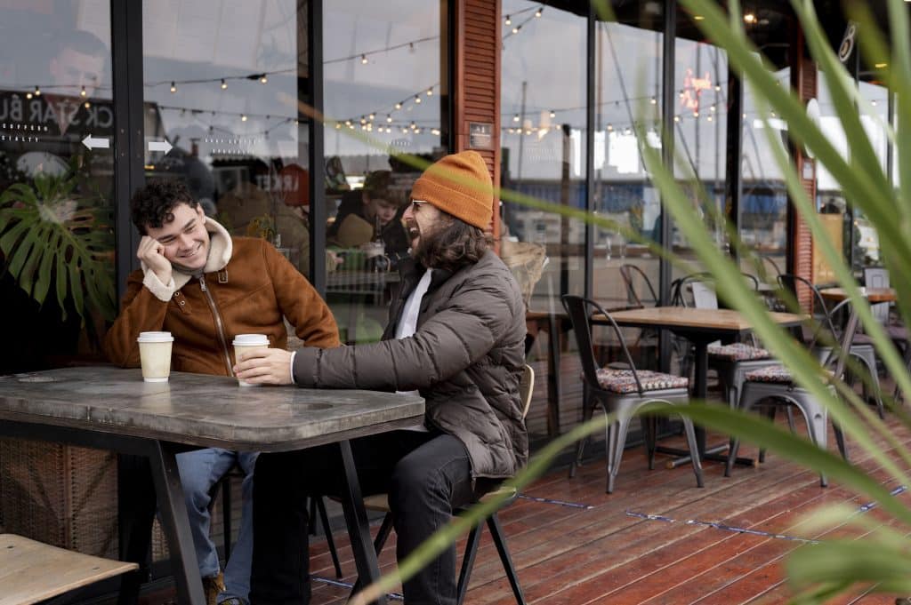 two male friends sitting with takeaway coffee cups outside a cafe and laughing together
