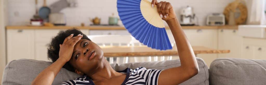 Woman fanning herself with a fan while lying back on a sofa with one hand to her forehead.