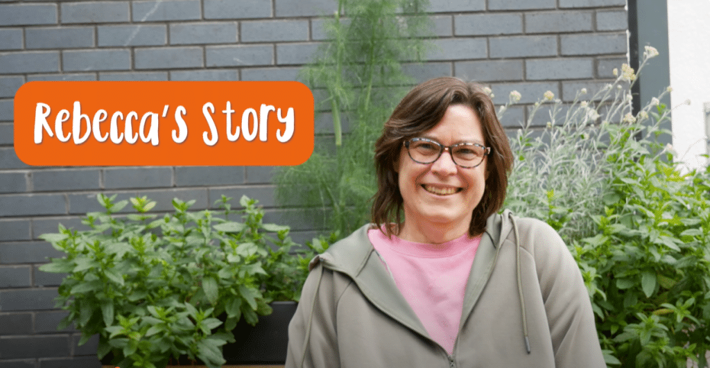 Rebecca sat in front of a brick wall and a planter of leafy green plants.