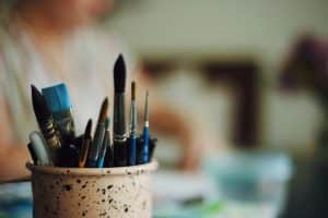A pot of paintbrushes sits on a desk in sharp focus, with an out of focus figure sat at the desk