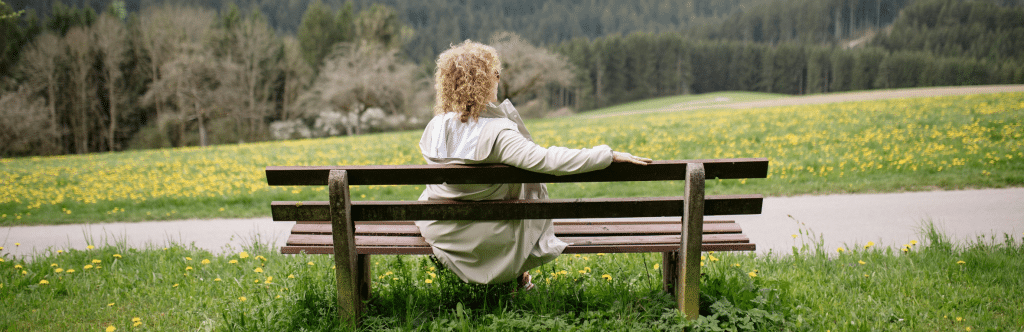 woman with curly blonde hair sitting on a park bench surrounded by grass and dandelions