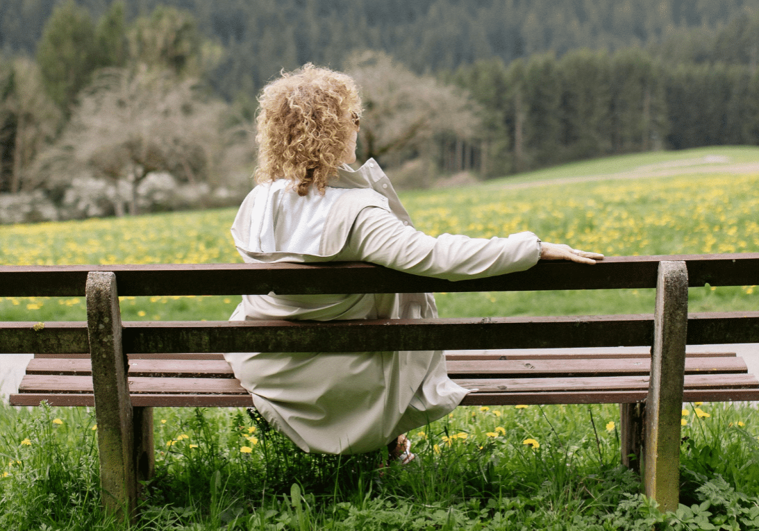 woman with curly blonde hair sitting on a park bench surrounded by grass and dandelions