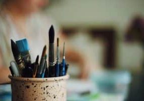 A pot of paintbrushes sits on a desk in sharp focus, with an out of focus figure sat at the desk