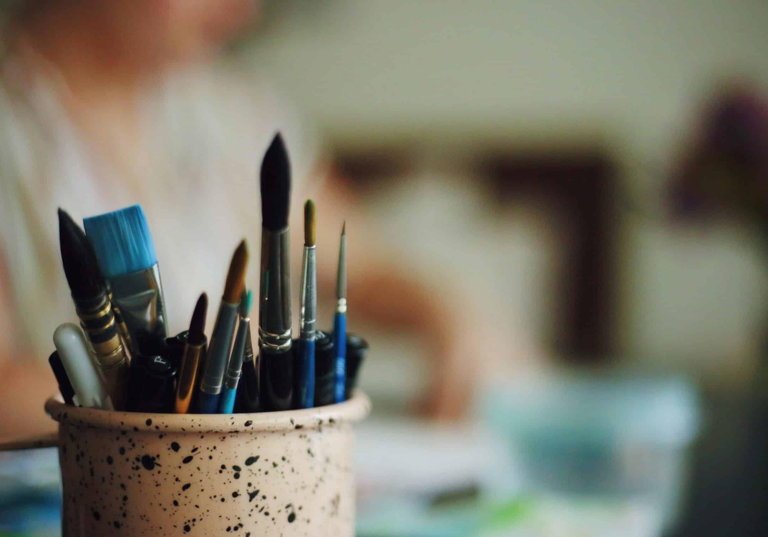 A pot of paintbrushes sits on a desk in sharp focus, with an out of focus figure sat at the desk